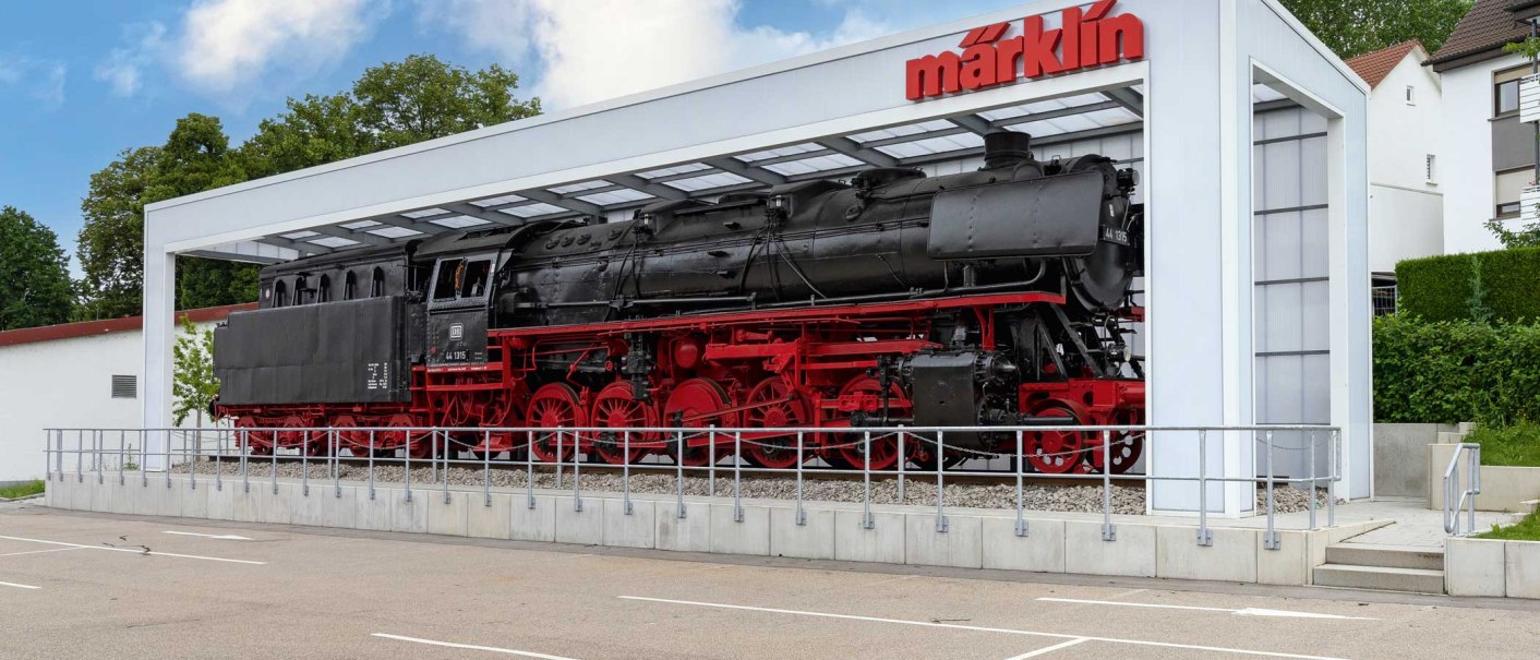 Dampflok der DR-Baureihe 44 unter einem modernen, überdachten Ausstellungsbereich mit Märklin-Logo. Der Himmel ist blau mit einigen Wolken., © Märklineum Dampflok der DR-Baureihe 44 unter einem modernen, überdachten Ausstellungsbereich mit Märklin-Logo. Der Himmel ist blau mit einigen Wolken., © Märklineum