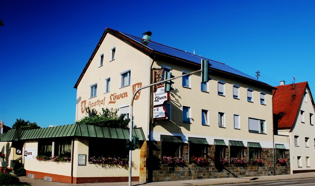 Der Gasthof Löwen mit beiger Fassade und roten Dächern, dekoriert mit Blumen, unter blauem Himmel., © Ulli Kellenbenz Der Gasthof Löwen mit beiger Fassade und roten Dächern, dekoriert mit Blumen, unter blauem Himmel., © Ulli Kellenbenz