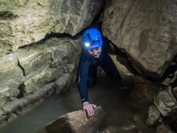 Eine Person mit Helm und Stirnlampe kriecht durch eine enge, wassergef&uuml;llte Passage in der Falkensteiner H&ouml;hle., &copy; SwabianTravel