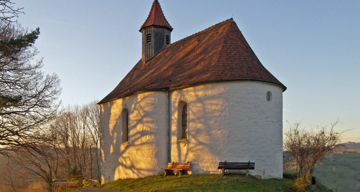 Die Marienkapelle in Wissgoldingen steht auf einem Hügel, beleuchtet von der Abendsonne. Bäume werfen Schatten auf die weiße Fassade., © Foto: Frieder Kopper Die Marienkapelle in Wissgoldingen steht auf einem Hügel, beleuchtet von der Abendsonne. Bäume werfen Schatten auf die weiße Fassade., © Foto: Frieder Kopper