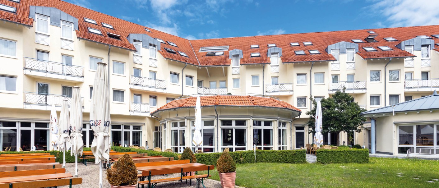 Hotel mit rotem Dach, Biergarten mit Holzbänken und geschlossenen Sonnenschirmen, umgeben von grüner Wiese und blühenden Sträuchern., © Seminaris Hotel Bad Boll Hotel mit rotem Dach, Biergarten mit Holzbänken und geschlossenen Sonnenschirmen, umgeben von grüner Wiese und blühenden Sträuchern., © Seminaris Hotel Bad Boll