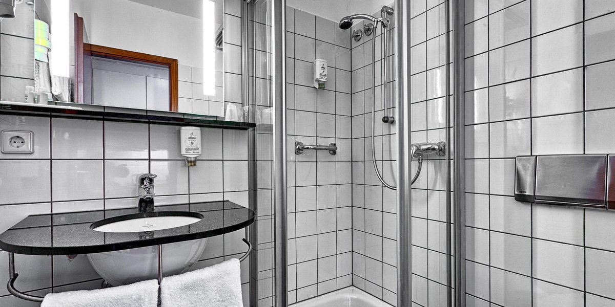 Modernes Badezimmer mit weiß gefliesten Wänden, Duschkabine, Waschbecken mit schwarzer Ablage und beleuchtetem Spiegel., © attimo