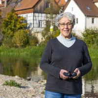 Eine &auml;ltere Frau mit Fernglas steht am Flussufer, im Hintergrund Fachwerkh&auml;user und herbstliche B&auml;ume., &copy; Stadt Besigheim