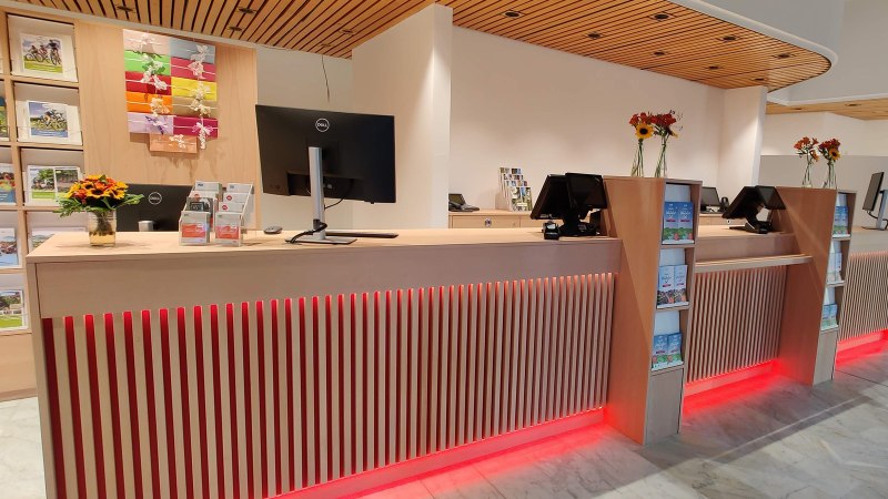 Moderner Empfangsbereich mit Holztheke, Computern, Broschüren und Blumenvasen. Helle Beleuchtung und stilvolle Einrichtung., © Larissa Philipp