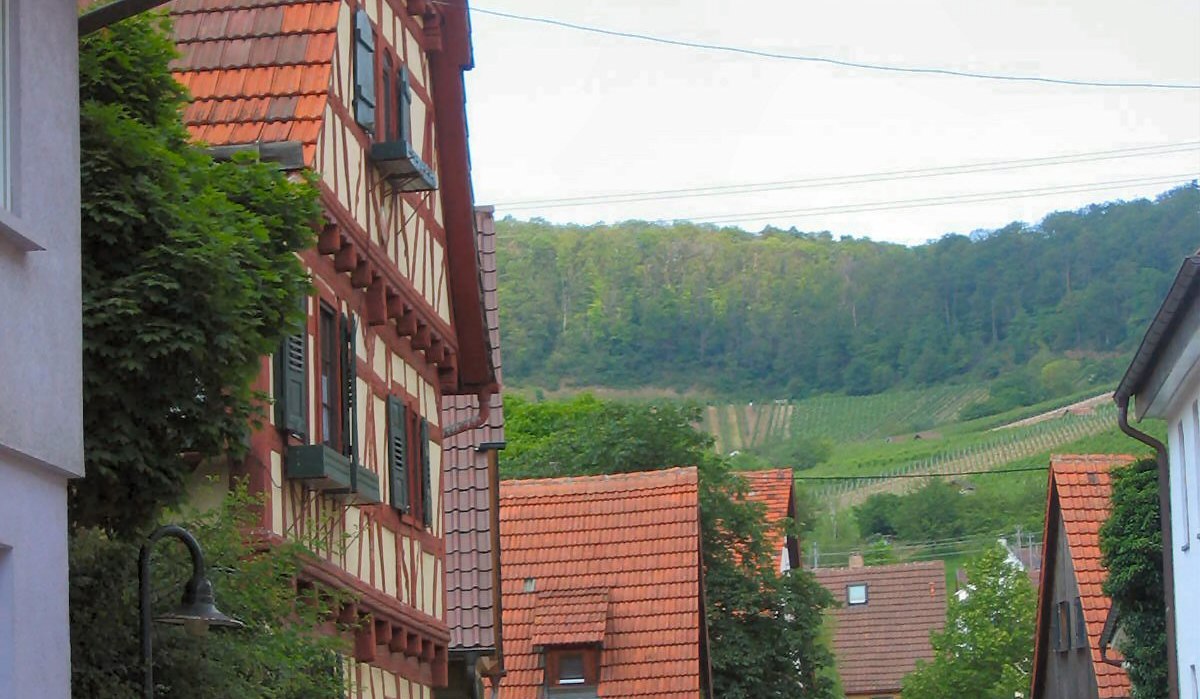 Fachwerkhäuser säumen eine ruhige Dorfstraße. Im Hintergrund sind grüne Weinberge zu sehen. Autos parken am Straßenrand., © Land der 1000 Hügel - Kraichgau-Stromberg Fachwerkhäuser säumen eine ruhige Dorfstraße. Im Hintergrund sind grüne Weinberge zu sehen. Autos parken am Straßenrand., © Land der 1000 Hügel - Kraichgau-Stromberg