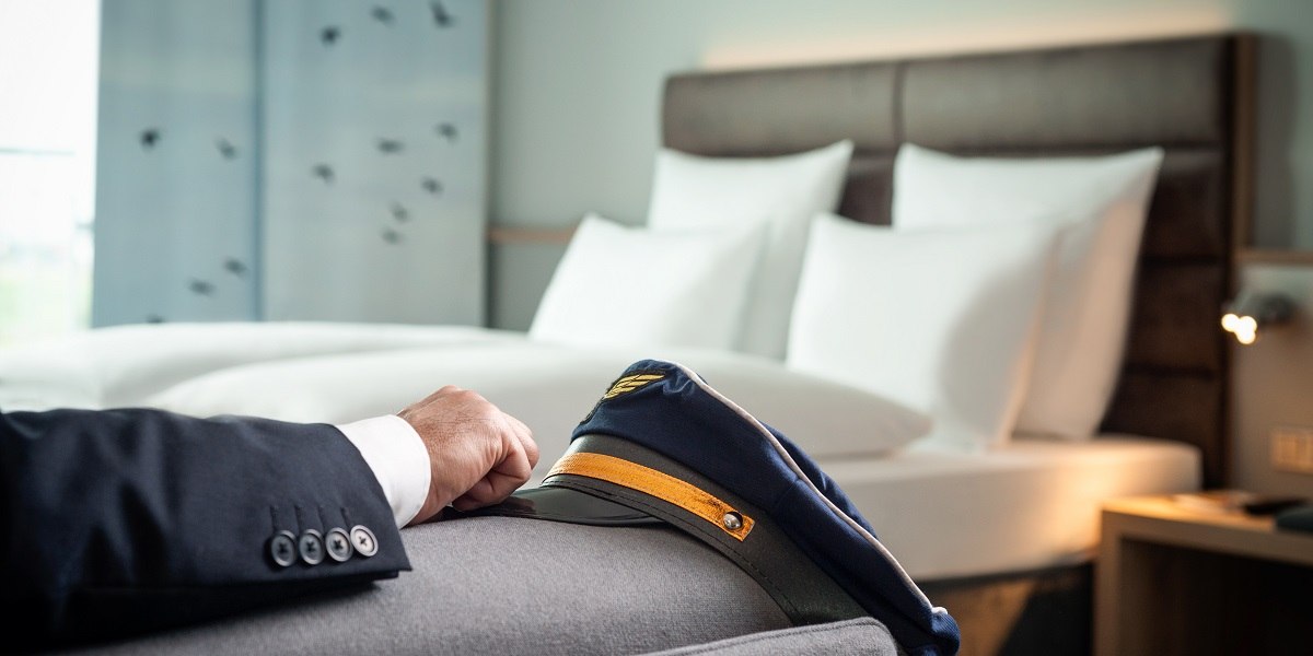 Ein Pilotensakko und -mütze liegen auf einem Bett in einem modernen Hotelzimmer mit weißen Kissen., © Essential by Dorint