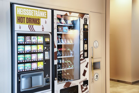 Ein Verkaufsautomat für Heißgetränke und Snacks steht in einem beleuchteten Innenraum. Links sind Getränke, rechts Snacks erhältlich., © B&B Hotel Stuttgart-Neckarhafen