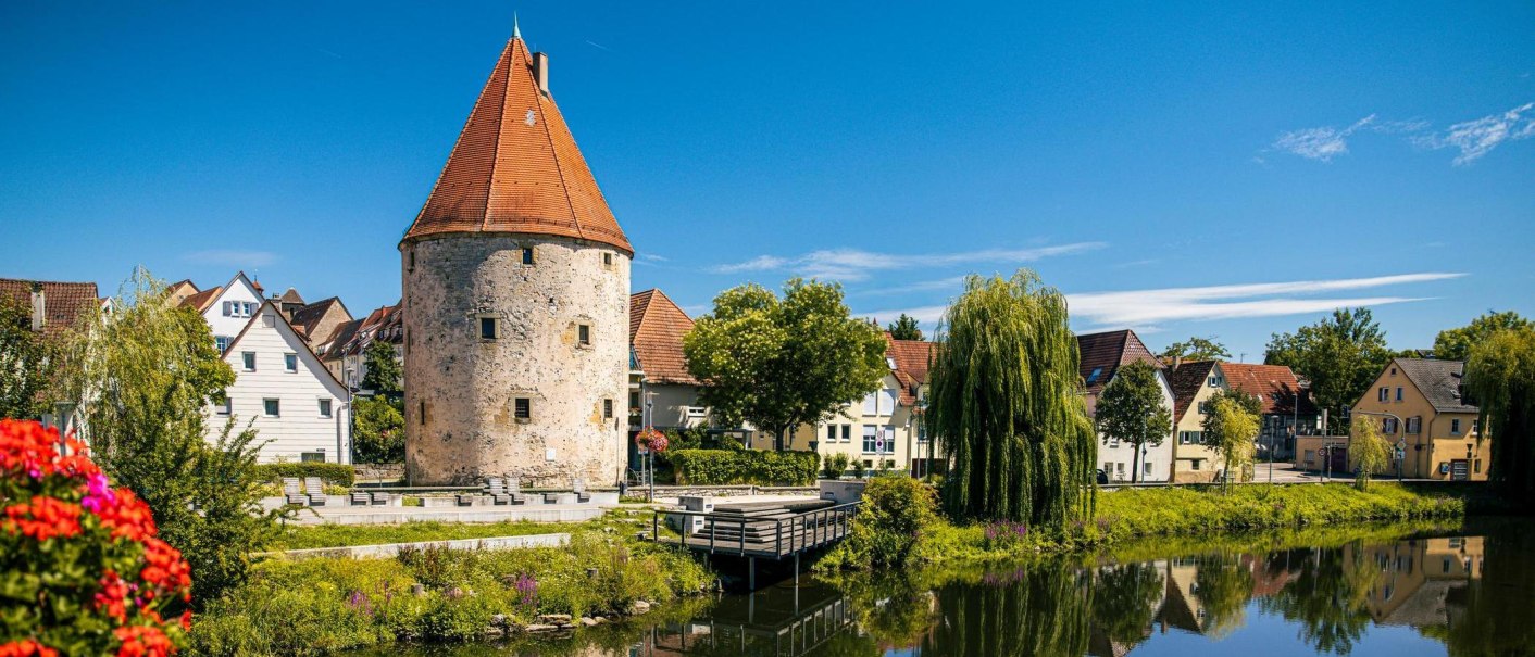 Der historische Pulverturm in Vaihingen an der Enz, umgeben von malerischen Häusern und einem ruhigen Fluss unter blauem Himmel., © SMG, Sarah Schmid Der historische Pulverturm in Vaihingen an der Enz, umgeben von malerischen Häusern und einem ruhigen Fluss unter blauem Himmel., © SMG, Sarah Schmid