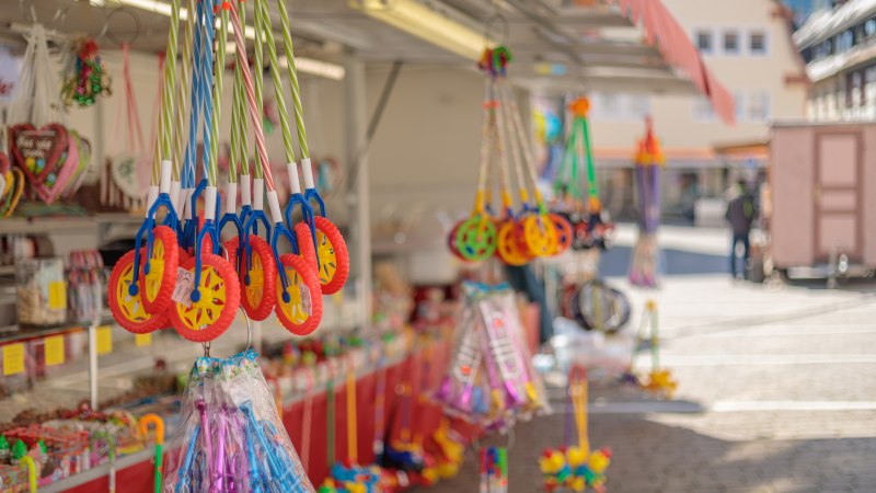 Verkaufsstand auf einem Krämermarkt mit buntem Spielzeug und Süßigkeiten. Im Hintergrund sind Marktstände und Gebäude zu sehen.