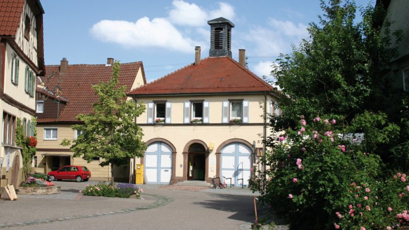 Das Rathaus in Ensingen mit rotem Ziegeldach und blauen Fensterläden, umgeben von blühenden Rosen und einem kleinen Platz., © Land der 1000 Hügel - Kraichgau-Stromberg Das Rathaus in Ensingen mit rotem Ziegeldach und blauen Fensterläden, umgeben von blühenden Rosen und einem kleinen Platz., © Land der 1000 Hügel - Kraichgau-Stromberg