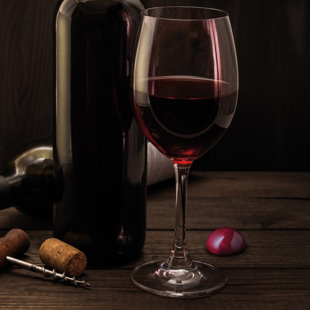 Ein Glas Rotwein steht neben einer Weinflasche auf einem Holztisch. Ein Korkenzieher und ein rosa Schokoladenbonbon liegen daneben., &copy; Mercedes-Benz AG