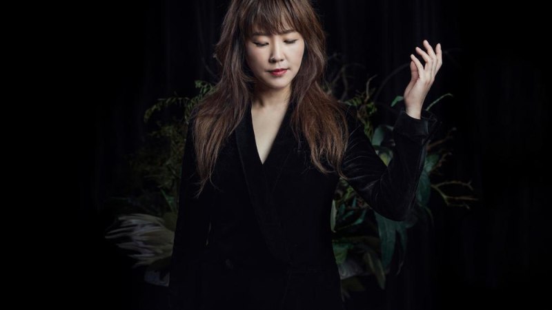 Eine Frau in schwarzer Kleidung steht vor einem dunklen Hintergrund mit Pflanzen. Sie hat die Augen geschlossen und hebt eine Hand leicht an., &copy; Youn Sun Nah &copy; Seung Yull Nah