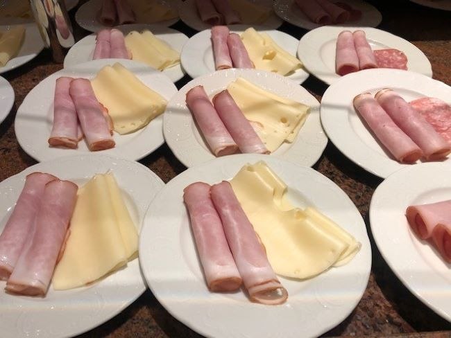 Weiße Teller mit aufgerolltem Schinken und Käsescheiben, auf einem Tisch arrangiert. Frühstücksvorbereitung., © Kronen Hotel GmbH