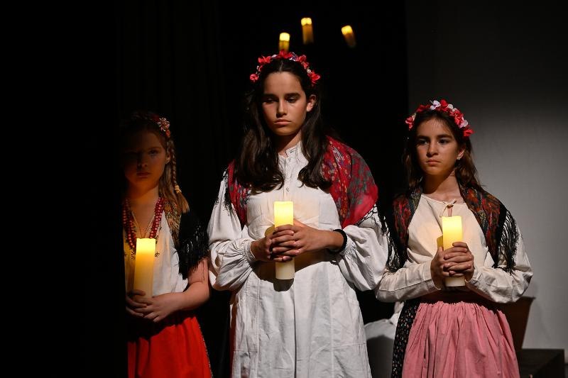 Drei Mädchen in traditioneller Kleidung mit Blumenkränzen halten Kerzen in einem dunklen Raum. Sie wirken konzentriert und ernst., © Kunstschule Labyrinth Drei Mädchen in traditioneller Kleidung mit Blumenkränzen halten Kerzen in einem dunklen Raum. Sie wirken konzentriert und ernst., © Kunstschule Labyrinth