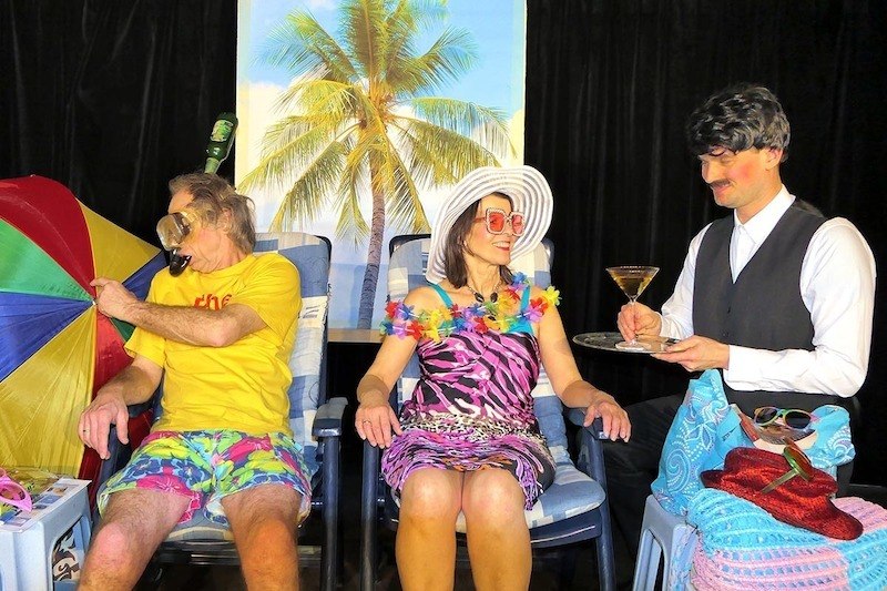 Ein Paar in bunter Urlaubskleidung sitzt vor einem Palmenhintergrund. Ein Kellner serviert ihnen einen Drink. Die Szene wirkt humorvoll und inszeniert., &copy; Galli Theater Backnang