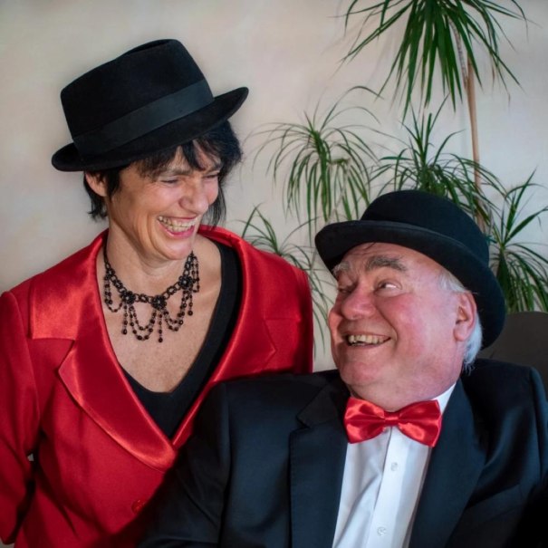 Ein Mann und eine Frau in eleganter Kleidung mit H&uuml;ten lachen herzlich. Die Frau tr&auml;gt einen roten Blazer, der Mann eine rote Fliege. Im Hintergrund sind Pflanzen zu sehen., &copy; Foto Susanne Schempp & Peter Appich
