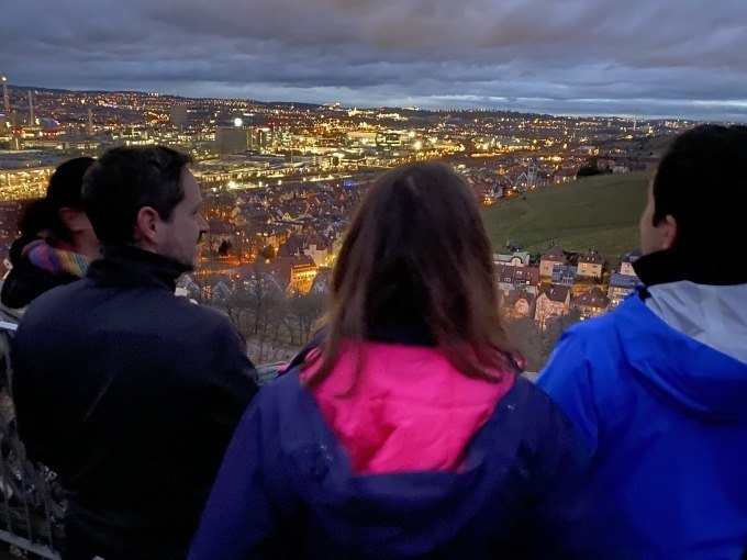 Vier Personen stehen auf einem Aussichtspunkt und blicken auf die beleuchtete Stadt Stuttgart bei D&auml;mmerung., &copy; Weingenuss Stuttgart