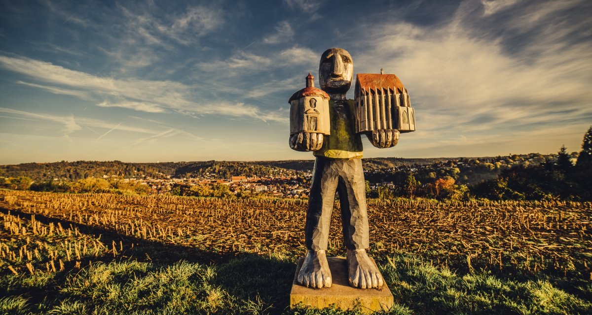 Eine Skulptur eines großen Wesens hält zwei Gebäude in den Händen, steht auf einem Feld unter einem weiten, bewölkten Himmel., © Landratsamt Ostalbkreis Eine Skulptur eines großen Wesens hält zwei Gebäude in den Händen, steht auf einem Feld unter einem weiten, bewölkten Himmel., © Landratsamt Ostalbkreis