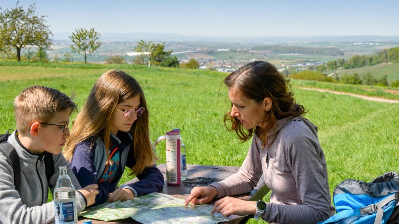 Drei Personen sitzen an einem Tisch im Freien und betrachten eine Karte. Im Hintergrund ist eine weite Landschaft mit Feldern und Bäumen zu sehen., © Stadtverwaltung Winnenden Drei Personen sitzen an einem Tisch im Freien und betrachten eine Karte. Im Hintergrund ist eine weite Landschaft mit Feldern und Bäumen zu sehen., © Stadtverwaltung Winnenden