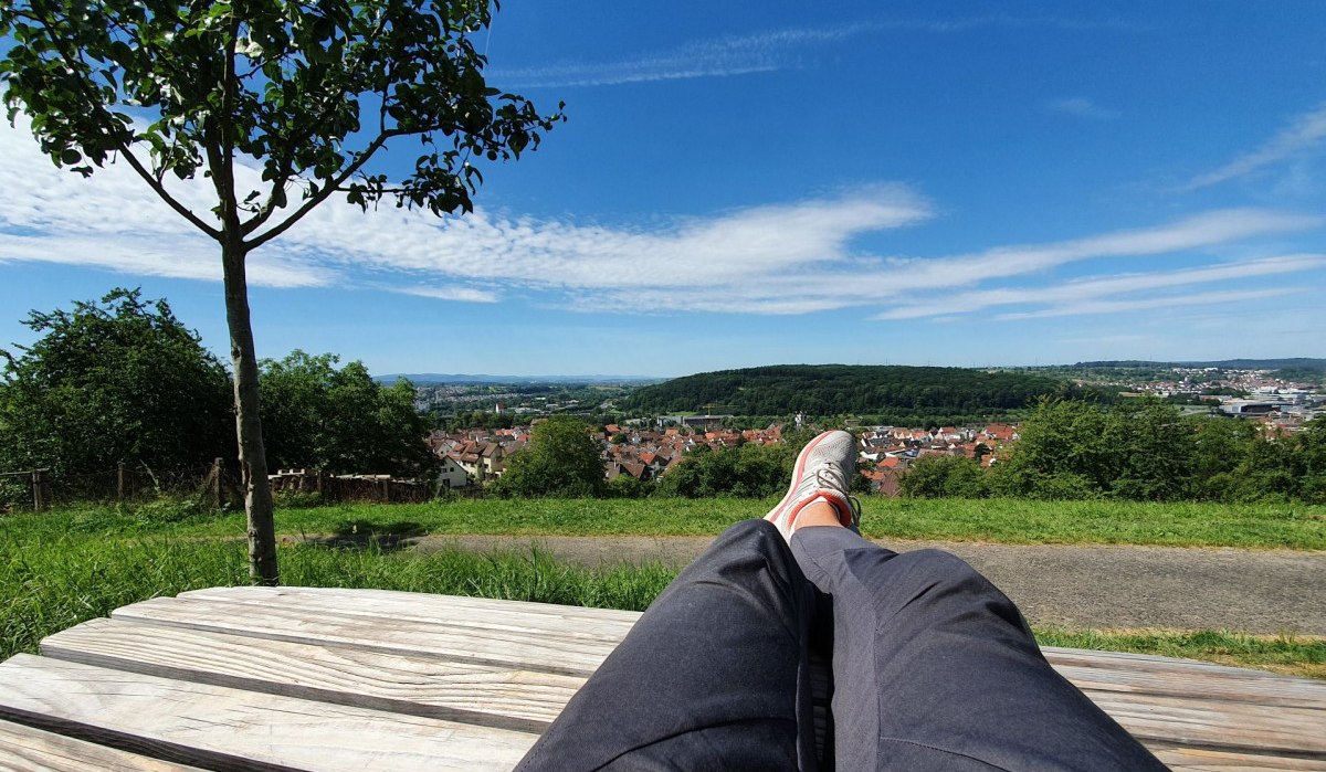 Person entspannt auf einer Holzbank mit Blick auf eine Stadtlandschaft und grüne Hügel unter blauem Himmel.