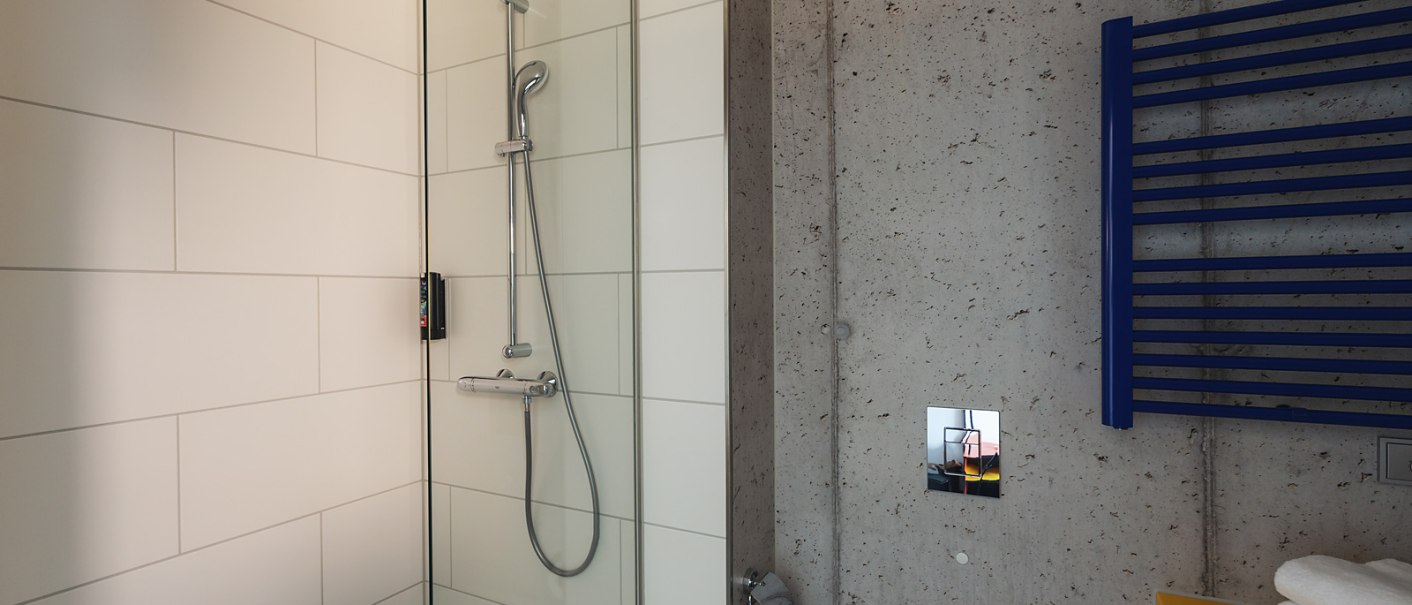 Modernes Badezimmer mit einer Glasdusche, weißen Fliesen, einem blauen Handtuchhalter und einer gelben Ablage mit Handtüchern., © the niu Form - Novum Hospitality