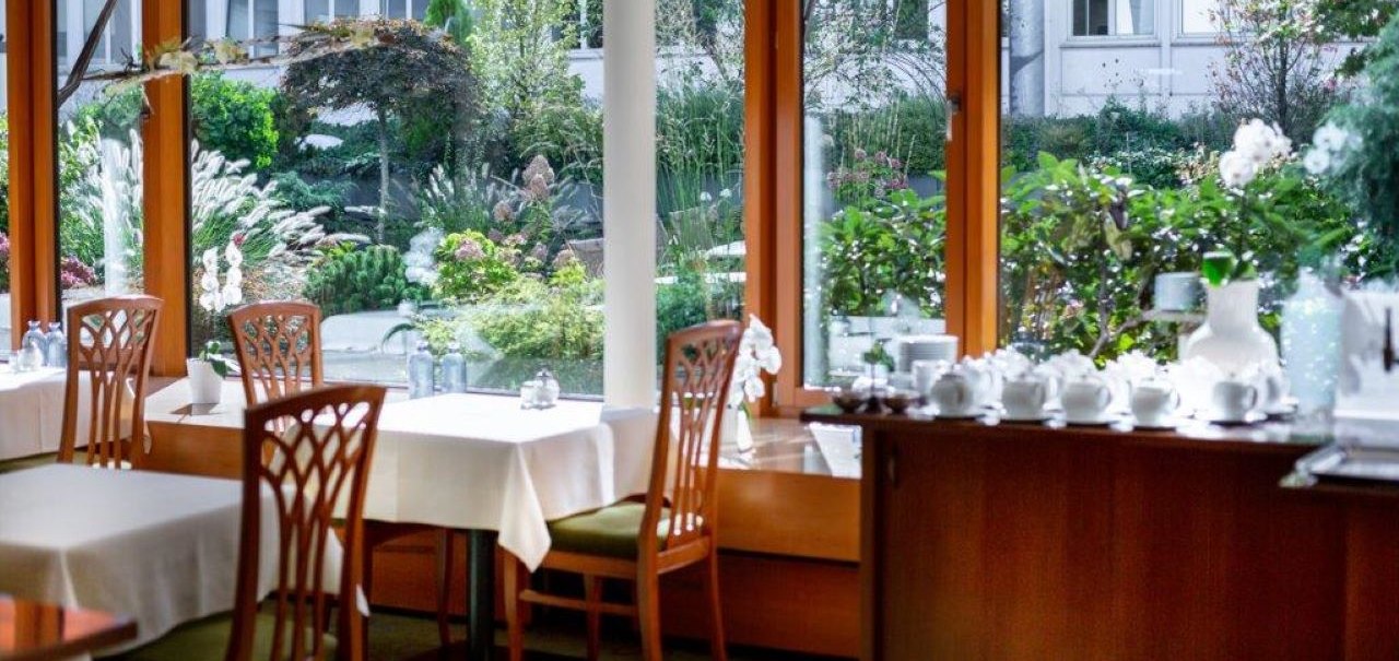Heller Frühstücksraum mit Holzmöbeln, weißen Tischdecken und Blick auf einen grünen Garten durch große Fenster., © Kronen Hotel GmbH Heller Frühstücksraum mit Holzmöbeln, weißen Tischdecken und Blick auf einen grünen Garten durch große Fenster., © Kronen Hotel GmbH