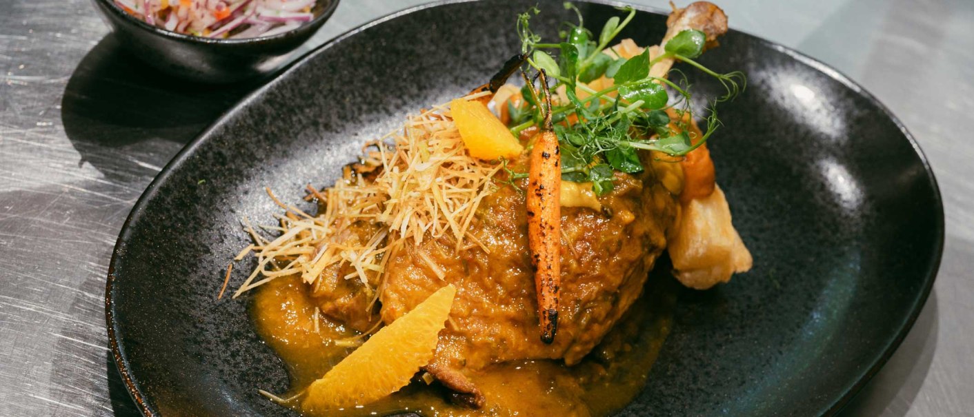 Ein kunstvoll angerichtetes Gericht auf einem schwarzen Teller, garniert mit Gemüse, Kräutern und einer Sauce. Im Hintergrund eine Schüssel mit Salat., © Kati Guenter