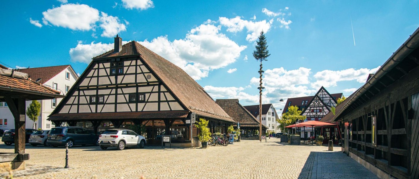Fachwerkhäuser am Kelterplatz in Metzingen unter blauem Himmel mit weißen Wolken. Autos parken auf dem gepflasterten Platz., © Stuttgart-Marketing GmbH, Sarah Schmid Fachwerkhäuser am Kelterplatz in Metzingen unter blauem Himmel mit weißen Wolken. Autos parken auf dem gepflasterten Platz., © Stuttgart-Marketing GmbH, Sarah Schmid