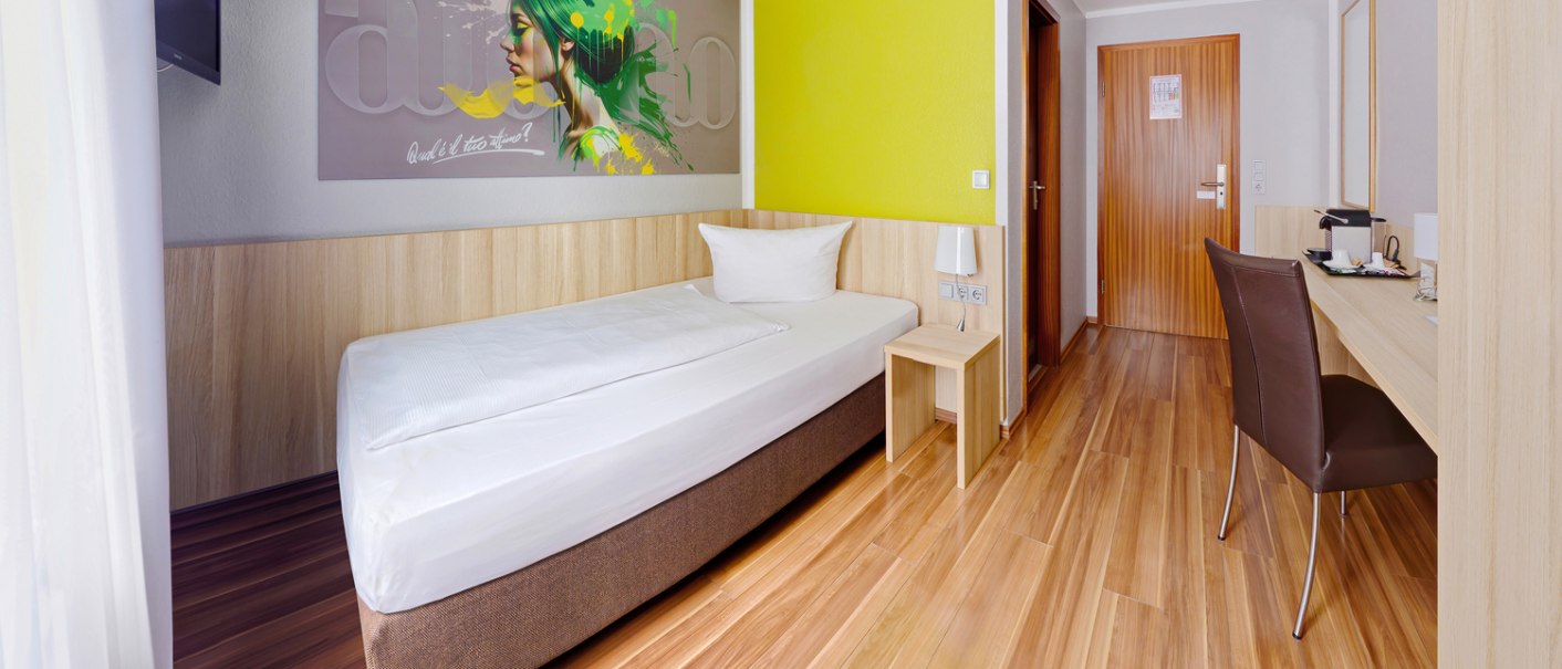 Modernes Einzelzimmer mit Holzboden, Einzelbett, Schreibtisch, Wandkunst und gelber Akzentwand., © attimo Hotel Stuttgart