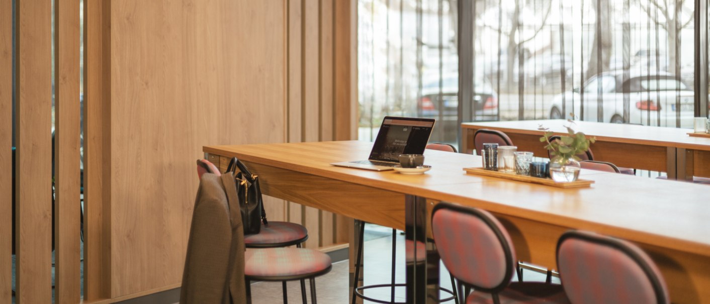 Moderner Frühstücksraum mit Holztischen, Stühlen und einem Laptop. Eine Jacke hängt über einem Stuhl, draußen sind Autos sichtbar., © Novum Management GmbH Moderner Frühstücksraum mit Holztischen, Stühlen und einem Laptop. Eine Jacke hängt über einem Stuhl, draußen sind Autos sichtbar., © Novum Management GmbH