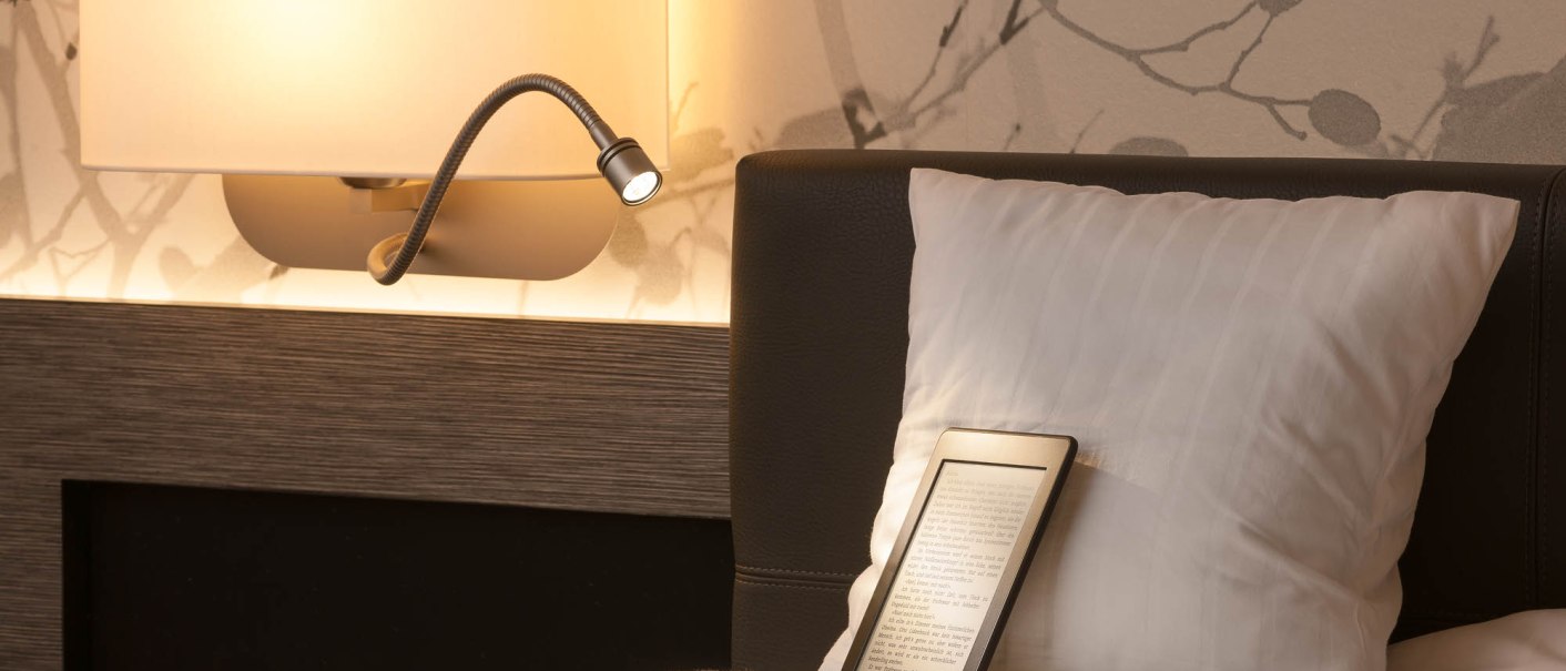 Ein Hotelzimmer mit einer Leselampe an der Wand, einem Kissen und einem E-Reader auf dem Bett. Die Tapete hat ein dezentes Muster., © MASEVEN Stuttgart SI-Centrum Ein Hotelzimmer mit einer Leselampe an der Wand, einem Kissen und einem E-Reader auf dem Bett. Die Tapete hat ein dezentes Muster., © MASEVEN Stuttgart SI-Centrum