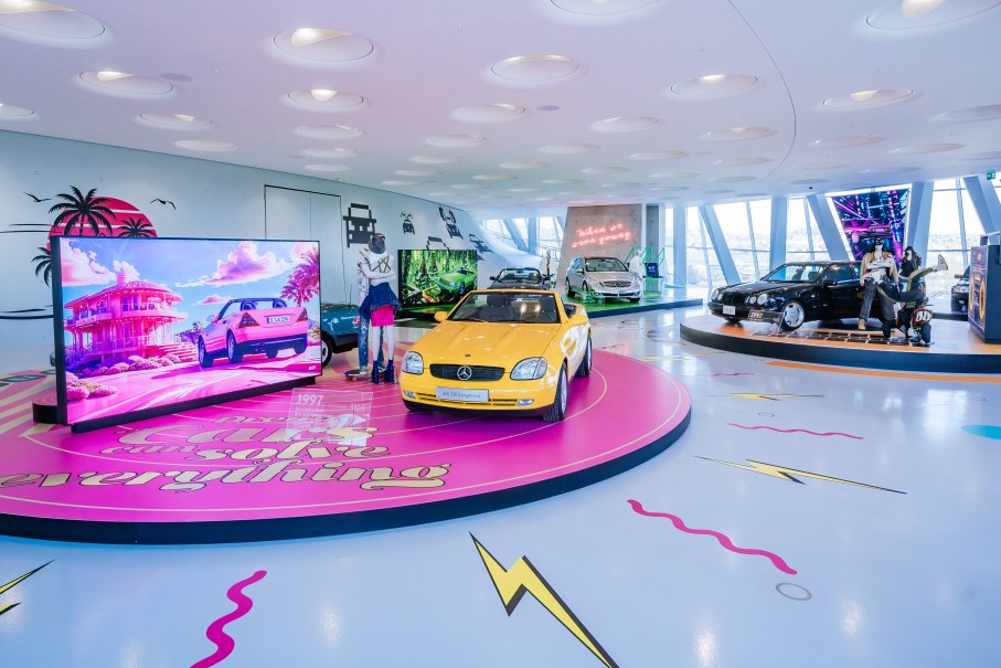 Bunte Ausstellung mit Youngtimer-Autos, darunter ein gelber Mercedes SLK. Neonlichter und Bildschirme mit Retro-Designs schmücken den Raum., © © Mercedes-Benz AG | Foto: Thomas Niedermüller