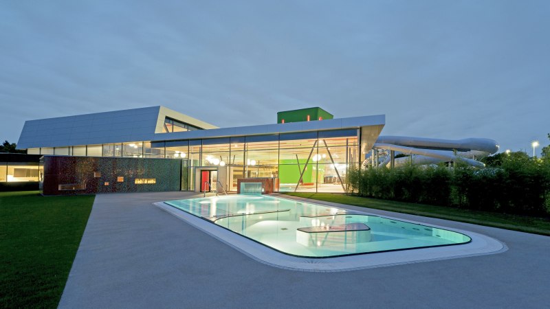 Modernes Schwimmbad mit beleuchtetem Außenpool und Glasfassade bei Dämmerung., © David Matthiessen Fotografie
