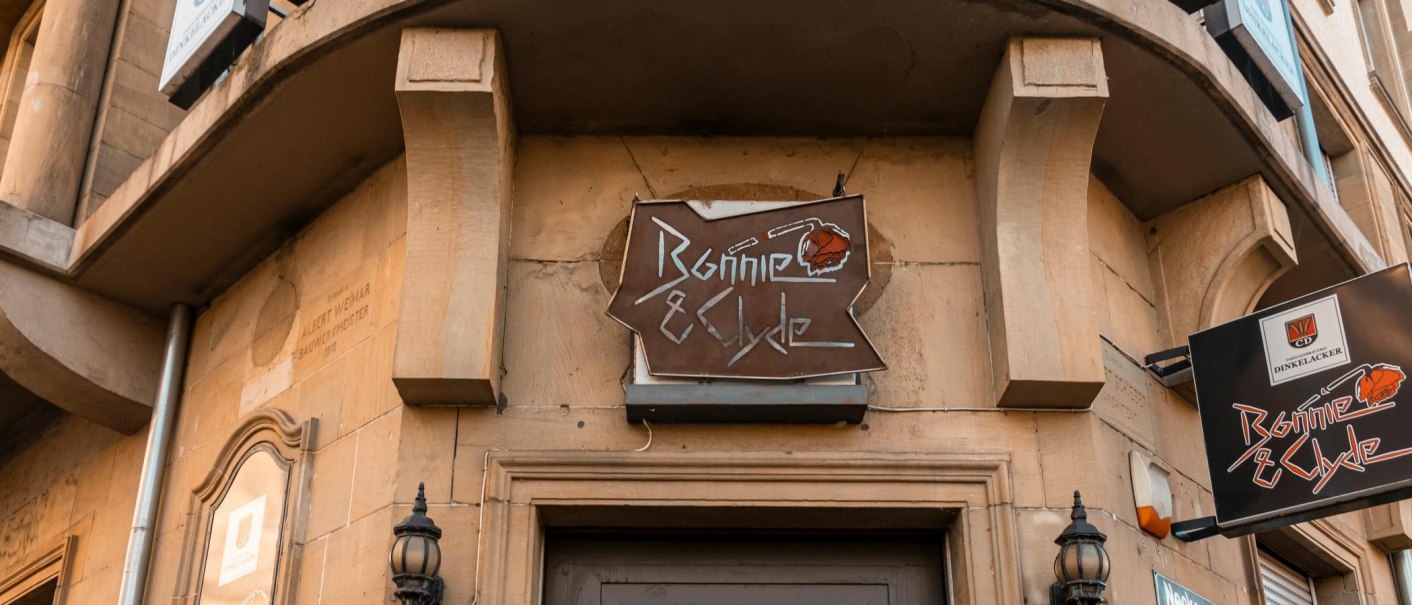 Ein Schild mit der Aufschrift 'Bonnie & Clyde' hängt an einem Gebäude. Die Fassade ist aus Stein, und es gibt dekorative Laternen., © SMG. Sarah Schmid Ein Schild mit der Aufschrift 'Bonnie & Clyde' hängt an einem Gebäude. Die Fassade ist aus Stein, und es gibt dekorative Laternen., © SMG. Sarah Schmid