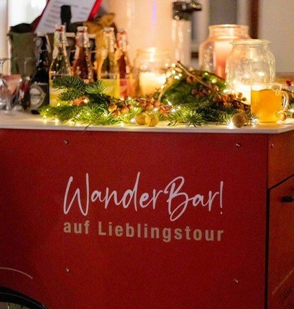 Ein mobiler Getr&auml;nkestand namens 'WanderBar!' ist festlich mit Lichtern und Tannenzweigen dekoriert. Verschiedene Getr&auml;nke sind auf dem Stand platziert.