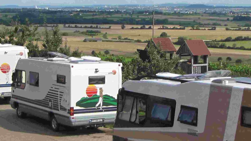 Wohnmobile parken auf einem Stellplatz mit Blick auf eine weite Landschaft und Felder. Im Hintergrund sind H&uuml;gel und eine Stadt zu sehen., &copy; PANORAMAWEINGUT Baumg&auml;rtner