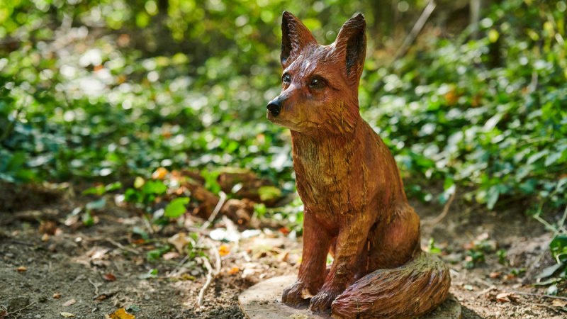 Eine detailreiche Holzskulptur eines Fuchses sitzt auf einem Erlebnispfad im Wald, umgeben von Laub und Pflanzen., © Natur.Nah. Schönbuch & Heckengäu Eine detailreiche Holzskulptur eines Fuchses sitzt auf einem Erlebnispfad im Wald, umgeben von Laub und Pflanzen., © Natur.Nah. Schönbuch & Heckengäu