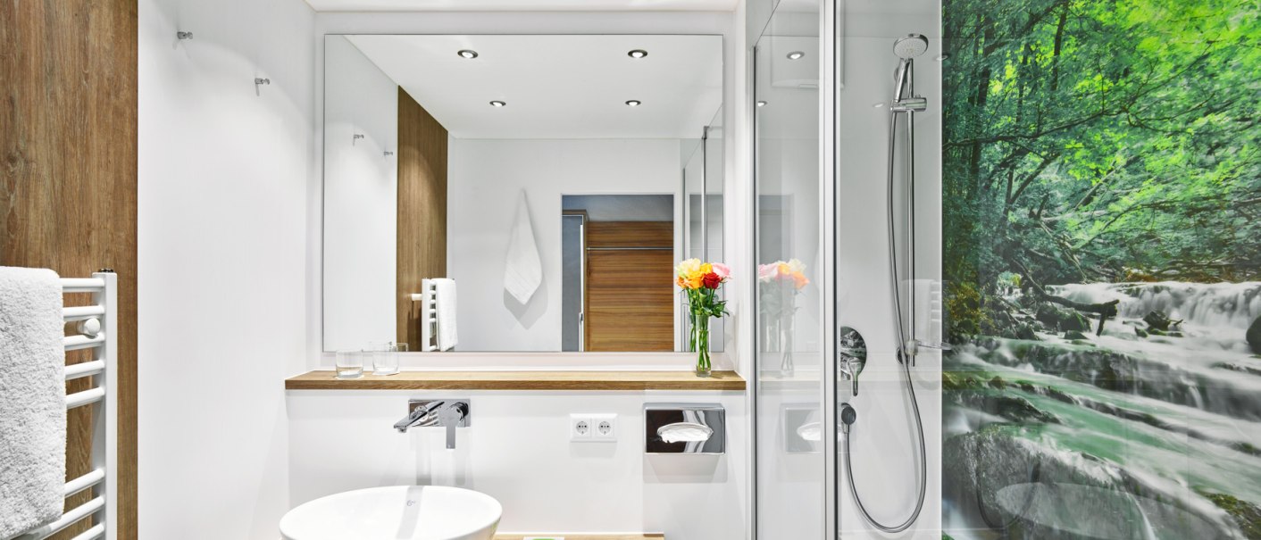 Modernes Badezimmer mit Holz- und Weißtönen, Waschbecken, Spiegel, Dusche und Wandbild eines Waldes., © Hotel Restaurant Lamm