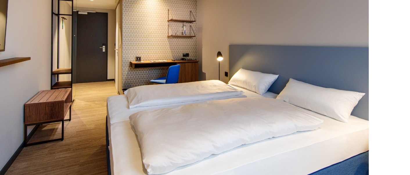 Modernes Hotelzimmer mit Doppelbett, Schreibtisch, blauer Stuhl und stilvoller Beleuchtung. Dezente Tapete und Holzmöbel ergänzen das Design., © Arthotel ANA Momentum