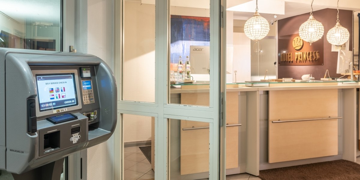 Hotelrezeption mit Selbstbedienungs-Check-in-Terminal und beleuchtetem Empfangstresen. Im Hintergrund das Schild 'Hotel Princess'., © Hotel Princess