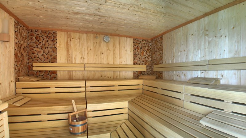 Innenansicht einer Sauna aus hellem Holz mit Bänken, einem Holzeimer und gestapeltem Holz an den Wänden., © TOMAS
