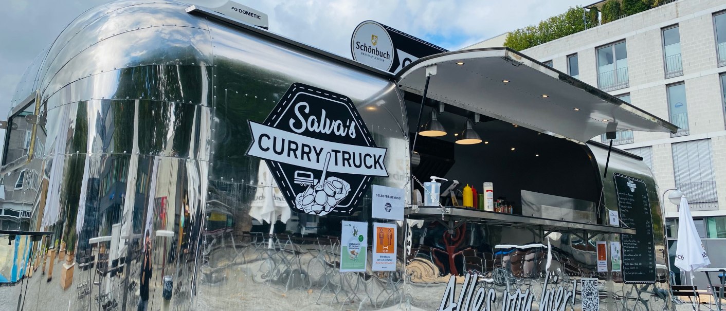 Ein glänzender Foodtruck mit der Aufschrift 'Salva's Curry Truck' steht im Freien vor einem modernen Gebäude., © Salva's Curry-Truck