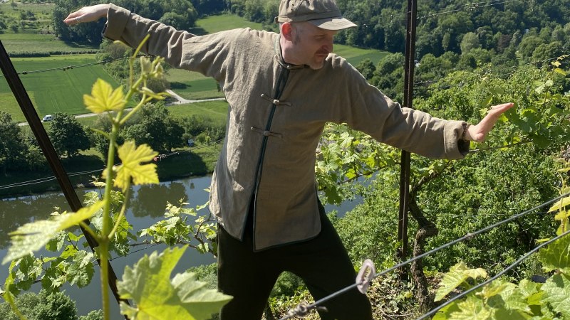 Person übt Qigong in einem Weinberg über dem Neckar. Im Hintergrund sind grüne Hügel und ein Fluss zu sehen. Sonniger Tag., © Karl Schüle und Daniel Steinbauer Person übt Qigong in einem Weinberg über dem Neckar. Im Hintergrund sind grüne Hügel und ein Fluss zu sehen. Sonniger Tag., © Karl Schüle und Daniel Steinbauer
