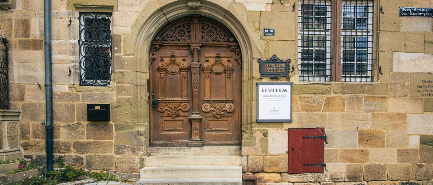 Eingang zur Sektkellerei Kessler in Esslingen. Eine kunstvoll geschnitzte Holztür wird von Schildern flankiert. Historische Architektur mit Details., © Stuttgart-Marketing GmbH, Sarah Schmid Eingang zur Sektkellerei Kessler in Esslingen. Eine kunstvoll geschnitzte Holztür wird von Schildern flankiert. Historische Architektur mit Details., © Stuttgart-Marketing GmbH, Sarah Schmid
