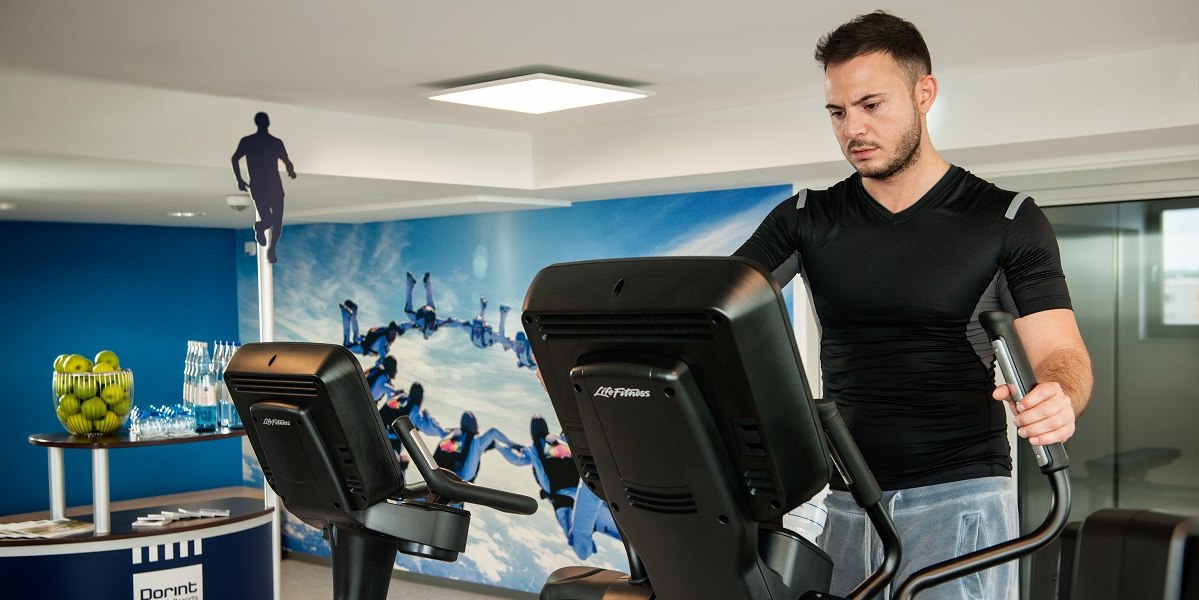 Ein Mann trainiert auf einem Crosstrainer in einem Fitnessraum. Im Hintergrund ist eine blaue Wand mit einem Fallschirmspringer-Motiv., © Essential by Dorint