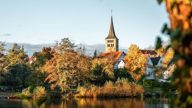 Eine malerische Kirche erhebt sich &uuml;ber herbstlich gef&auml;rbte B&auml;ume am Ufer eines ruhigen Sees, beleuchtet von warmem Sonnenlicht., &copy; Wirtschaftsf&ouml;rderung Sindelfingen GmbH