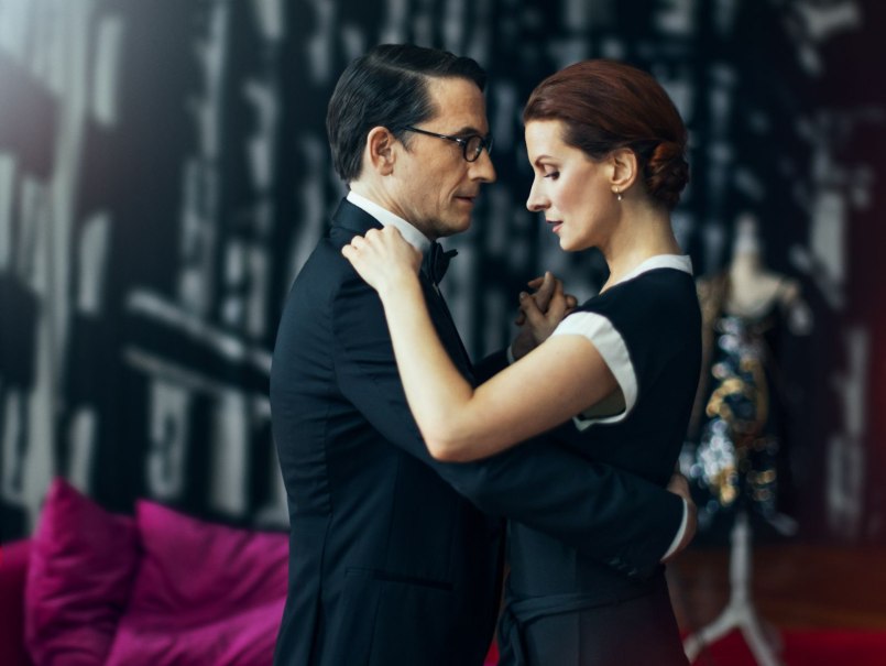 Ein Paar tanzt eng umschlungen in einem eleganten Raum mit dunklem Hintergrund und pinkem Sofa.