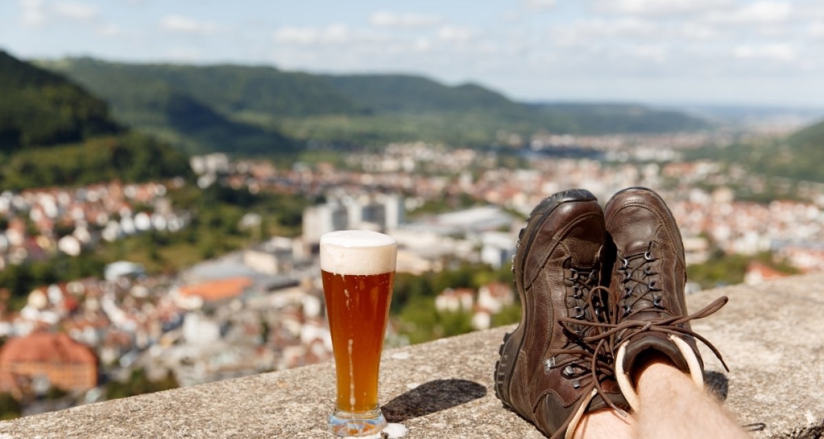 Wanderschuhe ruhen auf einer Mauer neben einem Glas Bier. Im Hintergrund ist die Stadt Geislingen und eine h&uuml;gelige Landschaft zu sehen., &copy; Landkreis G&ouml;ppingen
