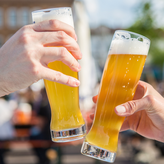 Zwei Personen sto&szlig;en mit gef&uuml;llten Weizengl&auml;sern an, im Hintergrund ein sonniger Biergarten., &copy; Ludwigsburg
