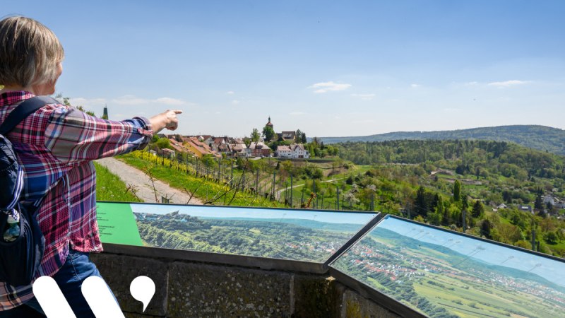 Eine Person zeigt auf eine malerische Landschaft mit Hügeln und einem Dorf. Im Vordergrund sind Informationstafeln zu sehen. Eine Person zeigt auf eine malerische Landschaft mit Hügeln und einem Dorf. Im Vordergrund sind Informationstafeln zu sehen.